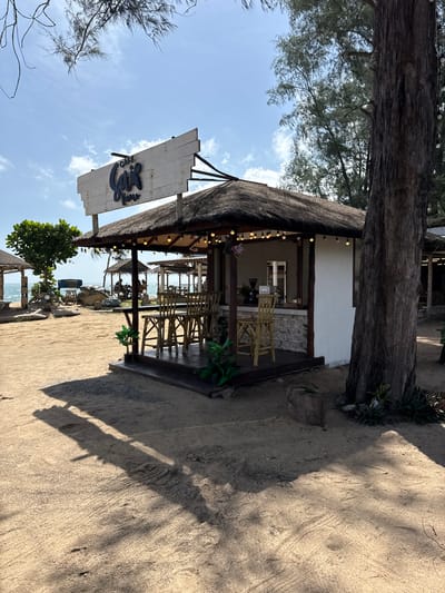 This image depicts an outdoor cafe at a beach in Ban Tha Chatchai, Thailand. The cafe features a thatched roof and a wooden deck with bamboo chairs and tables. A sign above the cafe reads "Cafe Gaja". The cafe is situated on a sandy beach with palm trees and other vegetation surrounding it. In the background, the ocean is visible, with other small structures that appear to be part of the beachfront establishment. The lighting suggests it is daytime with clear skies. There are no people prominently featured in the foreground, but silhouettes of individuals can be seen in the background at other seating areas. The overall scene conveys a relaxed, tropical atmosphere.