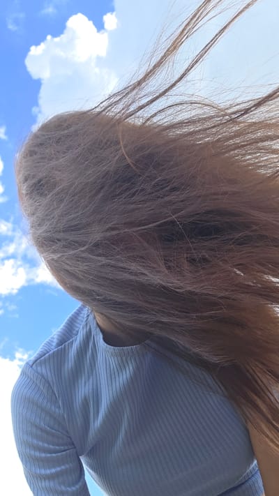 A low-angle, close-up shot captures a person's head and shoulders against a bright blue sky with scattered white clouds. The person's long, light brown hair is blowing across their face and obscuring it. They are wearing a light blue, ribbed, long-sleeved shirt. The scene appears to be outdoors.