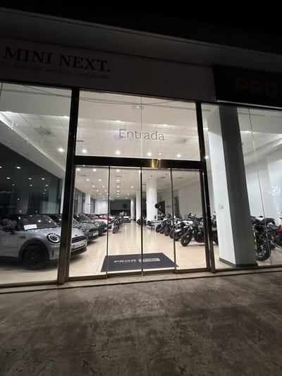 The image depicts the brightly lit exterior of a vehicle showroom at night, located in Polígon Industrial De Son Castello, Spain. The foreground features a dark, textured concrete pavement. The building's facade is composed of large, transparent glass panels framed by dark metallic supports, serving as display windows and a central entrance.
Above the left showroom section, a sign reads "MINI NEXT. Tu MINI de ocasion, certificado" (Your used, certified MINI). Above the main entrance, the word "Entrada" (Entrance) is visible in white lettering on the glass. The entrance consists of automatic sliding glass doors. Immediately inside, a dark rectangular mat bearing "PRO MAX" in white lettering is on the floor. A partial "PRO MAX" sign is also visible on the dark facade above the right section of the showroom.
The interior, well-illuminated by numerous recessed ceiling lights, showcases vehicles on a light-colored tiled floor. The left side displays several Mini Cooper automobiles, including a silver/grey Mini Cooper convertible with black wheels and a visible license plate "6094 MBN." Other Mini Coopers in various colors are arranged behind it. The right side of the showroom displays multiple motorcycles, predominantly dark in color, arranged in rows. Several motorcycles feature golden-colored front forks. No individuals are present, and no actions or interactions are occurring within the scene.