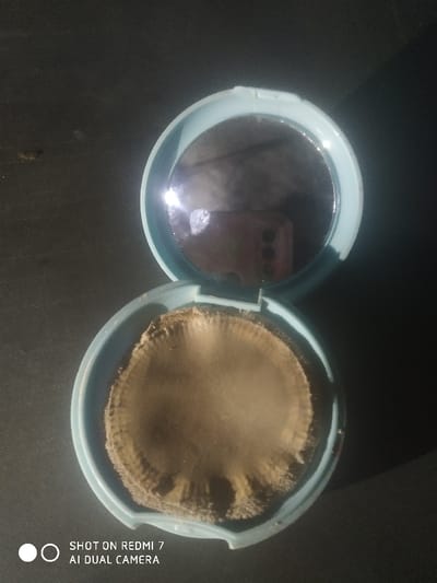 An open, light blue, circular cosmetic compact is positioned centrally on a dark, featureless surface. The compact's lid is open, revealing a circular mirror that reflects a portion of a dark-colored smartphone, including its camera lenses, indicating the device used to capture the image. The base of the compact contains a circular, light brown, textured material resembling a used makeup sponge or pressed powder, exhibiting wear and an uneven, slightly rippled surface texture. White text "SHOT ON REDMI 7 AI DUAL CAMERA" is visible at the bottom left. No specific location within Lamajang, Indonesia is discernible from the image content.