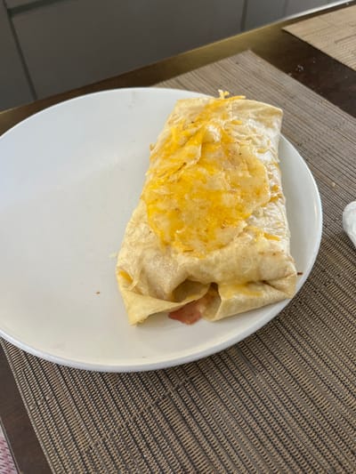 The image displays a single, large food item resembling a burrito or wrap, positioned on a white circular plate. The food item is oblong, wrapped in a light brown tortilla, and generously topped with melted, shredded yellow-orange cheese. Portions of a pinkish, cooked meat, consistent with ham or bacon, are visible at the bottom fold of the tortilla where it meets the plate. The white plate rests on a rectangular placemat featuring a woven brown and beige pattern. Behind the placemat, a dark wooden or laminate surface is visible, likely a table. In the upper background, a dark grey or black surface, possibly a wall or cabinet, is present, separated from the table by a narrow horizontal strip of a lighter, golden-brown material. No people are visible, and no discernible actions or interactions are taking place. The specific location within the city of San José Pinula, Guatemala, cannot be determined from the visual information presented in the image.