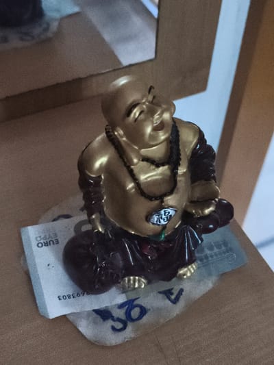 A golden, seated figurine of a laughing Buddha rests on a Euro banknote on a wooden surface. The figurine is dark brown from the waist down, depicting robes, and features a gold-colored, smiling face and torso. It wears a dark beaded necklace with a round pendant displaying a Chinese character. The Euro banknote, partially visible beneath the figurine, has text that reads "EURO EXPO" and a serial number that begins with "9693803". The background consists of a blurred wooden shelf and a light blue wall. The scene appears to be indoors, possibly in a home or shop. No specific location within Eitorf, Germany can be determined from the image.