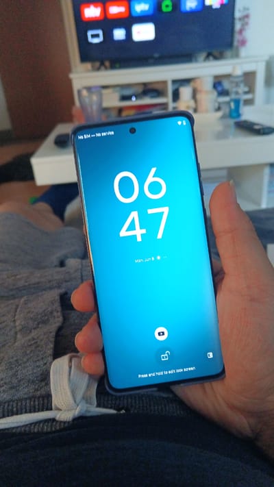 A person is holding a smartphone with a blue screen showing the time "06:47" and the date "Mon, Jun 8". The top of the screen displays "No SIM - No service". At the bottom of the screen, there are icons for a camera and a lock, with the text "Press and hold to edit lock screen". The background shows a television with streaming service logos, a shelf with various items, and a part of a sofa. The setting appears to be indoors, likely a living room. The image does not provide any information about the location in Ginsheim-Gustavsburg, Germany.
