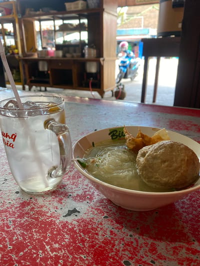 A close-up shot shows a glass of iced drink next to a bowl of noodles and meatballs on a distressed red table. The glass is clear with a handle and a straw, filled with ice, water, and a slice of lemon. Red lettering is visible on the glass. The bowl contains a broth with noodles, a large meatball, and some fried wontons or dumplings. In the background, a blurry scene depicts a street with a motorcycle and a person wearing a pink helmet. The environment appears to be an outdoor eatery or roadside stall, possibly in Sungai Sipai, Indonesia. The lighting suggests it is daytime.