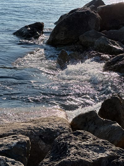 A close-up, sunlit view of waves crashing against large rocks on the coast of Alimos, Greece. The water is a deep blue, sparkling with highlights from the sun. White foam and spray erupt where the waves meet the rocks. The rocks themselves are varied in size and texture, with some appearing smooth and others rough and weathered. The scene captures the dynamic interaction between the sea and the rocky shoreline.