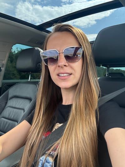 A woman with long, blonde hair and sunglasses is sitting in the passenger seat of a car, looking at the camera. The car has a sunroof, and the sky is visible through it, with white clouds against a blue background. The interior of the car is black leather. The woman is wearing a black t-shirt with a colorful graphic design. The scene appears to be in Troyanovo, Bulgaria, as indicated by the context.