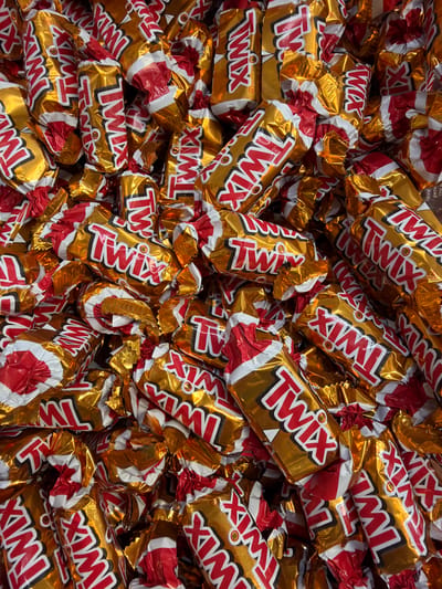 This is a close-up, overhead shot of a large pile of individually wrapped Twix candy bars. The wrappers are shiny gold with red and white lettering that says "Twix". The candy bars are oriented in various directions, creating a dense, overlapping mass. The lighting is bright and even, highlighting the reflective surfaces of the wrappers. There are no people visible in the image. The context of Sokilnyky, Ukraine is not apparent from the visual information provided.