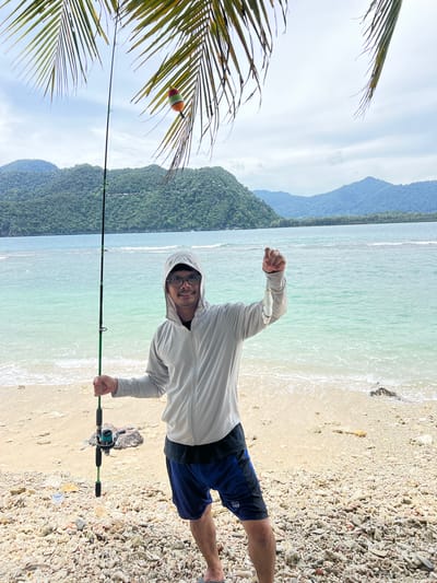 A man stands on a beach in Babahie, Indonesia, holding a fishing rod and a small fish. He is wearing a light-colored hooded jacket, dark shorts, and glasses, and he smiles at the camera. The beach is covered in small rocks and coral fragments. Behind him, the turquoise ocean meets a sandy shore, and in the distance, lush green mountains rise against a cloudy sky. Palm fronds hang from above, partially obscuring the scene. The fishing rod has a colorful float attached to its line. The man is holding the fish up for display.