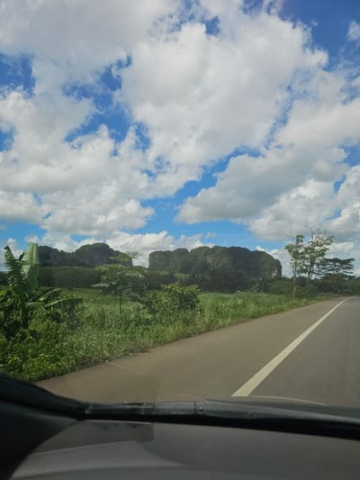 The image captures a scenic view from a vehicle on a paved road in Ban Nai Sa, Thailand. The road, marked with a white line, curves towards the right, disappearing into the distance. On the left side of the road, lush green vegetation thrives, including tall grasses, leafy plants, and banana trees. In the background, majestic, verdant karst mountains rise sharply, their sides covered in dense foliage. The sky above is a vibrant blue, dotted with large, fluffy white clouds, creating a picturesque and serene atmosphere. The perspective suggests the photo was taken from the driver's or a front passenger's seat, as the edge of the vehicle's dashboard is visible at the bottom of the frame.