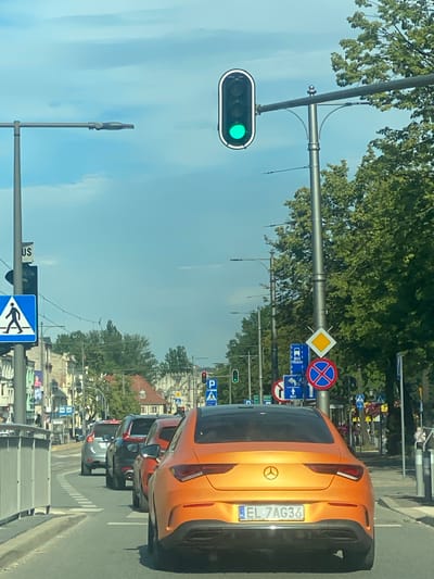 The image shows a street scene in Pabianice, Poland, from the perspective of someone in a vehicle. The sky is clear and blue, suggesting a sunny day. A traffic light with a green signal is prominently displayed on the left side of the frame. In the foreground, an orange car with the license plate "EL 7AG36" is visible, followed by other vehicles in traffic.
Several traffic signs are present, including a yield sign (a yellow diamond with a white border), a "no parking" sign (a blue circle with a red border and a red cross), and various bus signs. Some pedestrian crossing signs are also visible. Trees line the street, and buildings can be seen in the background. The overall impression is a typical urban street scene during daylight hours.