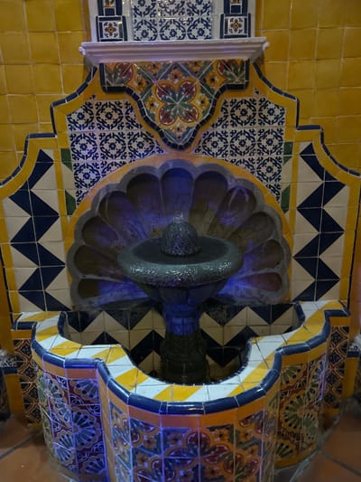 The image depicts a decorative tile fountain in Santa Ana Yancuitlalpan, Chilhuacán, Mexico. The fountain consists of a tiered stone basin with water illuminated by blue lights. The surrounding wall is adorned with yellow, blue, white, and orange patterned tiles in a geometric design.