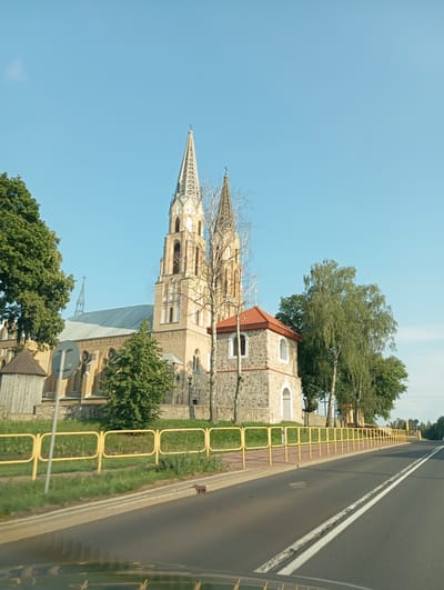 This is a full shot, exterior, eye-level, horizontal photograph taken on a sunny day of a church in the town of Korycin, Poland. The church is made of light brown brick and has two tall spires with crosses on top. A smaller stone building with a red tiled roof is attached to the church. The church is surrounded by trees and a yellow metal fence. A road runs in front of the church, with white dividing lines. The sky is clear blue. There are no people or animals visible in the image.