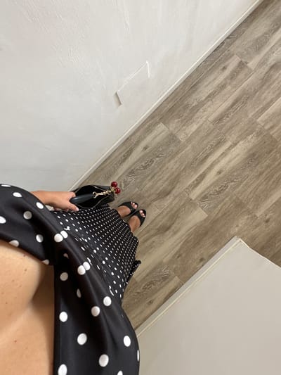 An overhead, slightly angled medium shot shows a person's torso, legs, and feet in a dimly lit interior space. The person is wearing a black dress with white polka dots and black open-toed sandals. A black handbag with a gold chain and cherry-shaped charms is held by the person's hand, resting on the dress. The floor is covered with light brown wood-patterned laminate flooring, laid diagonally. The wall to the left is painted white with a textured finish. The right side of the image shows the edge of a white baseboard and a sliver of another white surface, possibly a door or wall. The lighting appears to be artificial, casting subtle shadows. The image does not provide any specific visual cues to determine a precise location in Torremolinos, Spain, beyond it being an indoor setting.
