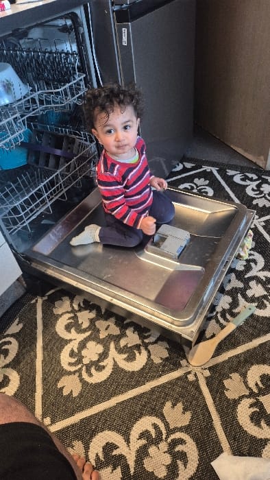 An indoor scene in Melle, Belgium, depicts a young child seated inside the lower rack of an open dishwasher. The child, with dark curly hair and light skin, wears a red, white, and dark blue striped long-sleeved shirt over a light green undershirt, dark blue pants, and light-colored socks. The child is positioned facing the camera, maintaining direct eye contact. The dishwasher's interior is stainless steel; its upper rack contains a white bowl with a floral pattern and a blue cup. The detergent dispenser is visible on the inner door panel where the child is seated. The floor is covered with a black and white patterned rug featuring floral and geometric motifs. To the right of the dishwasher, a wooden spatula with a light blue handle rests on the rug. A fragment of an adult's leg, with dark shorts and a bare foot, is visible in the lower-left foreground, suggesting the presence of an observer or photographer. The background consists of dark gray cabinetry or walls and a light brown vertical surface.
