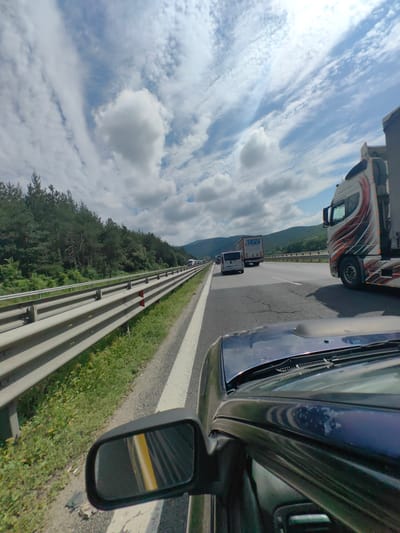 The media file captures a view from the driver's seat of a car traveling on a highway in Potop, Bulgaria. The scene is a highway with multiple lanes and traffic, including several trucks and vans. The highway is bordered by a metal guardrail and dense green trees on the left, with rolling hills in the background. The sky is filled with dramatic white and grey clouds against a bright blue background, suggesting a partly cloudy day.
The prominent subjects are the vehicles on the highway. A large, white truck with a stylized, colorful design on its side is in the foreground, on the right side of the frame. To its left, a white van is visible, and further down the road, another truck with "ZEPP TRANSPORT" written on its side is seen. The perspective suggests the car is moving at a moderate speed, indicated by the reflection of the road and yellow line in the side mirror.
The activity is simply traveling on the highway. There are no people clearly visible in the frame, but the presence of vehicles implies human activity related to transportation. The time of day appears to be daytime, given the bright sky and clear visibility. The weather seems pleasant, with a mix of sun and clouds. The lush green vegetation suggests a warmer season, likely spring or summer. The overall mood conveyed is one of travel and transit.
The text visible in the image includes "ZEPP TRANSPORT" on the side of one of the trucks. The location context of Potop, Bulgaria, is provided separately.