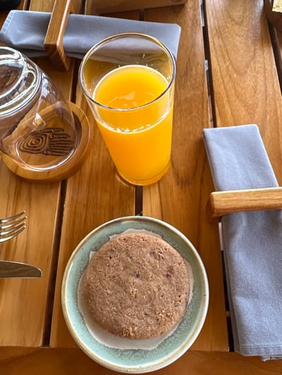 The scene depicts a tabletop setting in La Fortuna, Costa Rica. A light brown wooden slatted table serves as the primary surface. Centered in the upper portion is a clear glass containing orange juice. To its left, an overturned clear glass or carafe rests on a light brown wooden coaster adorned with a dark, stylized spiral pattern. Below the orange juice glass, positioned centrally at the bottom, is a single, round, light brown cookie on a small, round ceramic plate with a light blue-green rim. To the far left, a silver fork with four tines and a silver knife are partially visible. Two folded gray cloth napkins, each secured by a light brown wooden napkin ring, are present; one is in the upper right quadrant, and the other is partially visible in the lower right. The items are arranged consistent with a meal or snack service.