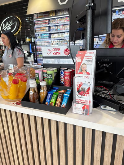 The image captures a scene inside a Lukoil service station in Chuchuligovo, Bulgaria. In the foreground, a counter displays a variety of beverages and snacks. To the left, a glass basin holds several small plastic bottles filled with a yellow liquid, likely juice, submerged in ice. Adjacent to this are several cans of Aloe Vera drinks, some labeled "Aloe Vera Original" and "Aloe Vera Pomegranate". Further to the right, a black display organizer holds an assortment of chocolate bars, including "Roshen Milk Chocolate", "Roshen Chocolate", and other unidentified brands. Behind the counter, a computer monitor is visible. To the left of the monitor, a woman with dark hair is standing, wearing a grey Lukoil branded shirt. To the right of the monitor, a woman with curly blonde hair is seated and looking down. Several promotional brochures and leaflets are displayed on a stand in front of the monitor. The overall environment suggests a retail setting within a gas station, focusing on impulse purchase items.