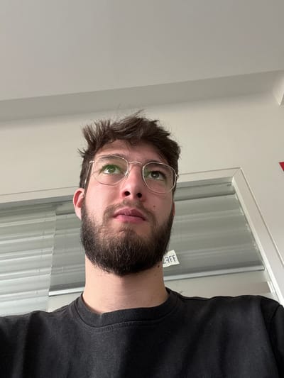 The image depicts a close-up, upward-angled shot of a young adult male located in Lièpvre, France. The subject has dark brown, disheveled hair and a full, dark brown beard. He is wearing gold-rimmed, hexagonal eyeglasses, and his eyes are directed upwards and slightly to the right of the frame, showing a faint green reflection in the lenses. He is wearing a dark-colored, possibly black, crew-neck t-shirt. The background consists of a light-colored, likely white or light grey, ceiling in the upper portion of the frame. Below this, a window or glass partition with horizontal blinds or slats is visible to the left and behind the subject. A small, white sticky note with the partially visible text "AFF" (likely part of "STAFF") in dark lettering is affixed to the glass. To the far right, a white door frame or partition edge is present, with a small portion of a red object, possibly a sign, visible in the upper right corner. The lighting is bright and even, illuminating the subject's face.