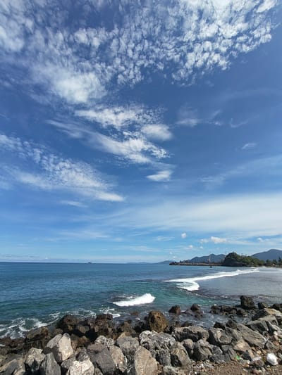 The scene is a coastal view in Naga Umbang, Indonesia. In the foreground, large, dark gray rocks form a barrier, with a small amount of trash visible amidst them. Waves are crashing against these rocks and the nearby shore. The water transitions from a dark blue further out to a clear, turquoise near the shore. A small sandy beach stretches along the right side of the frame, leading to a rocky outcrop and a constructed breakwater. Cranes are seen on the breakwater. Mountains are in the background, under a mostly blue sky with scattered white clouds. A small ship is visible in the distant water.