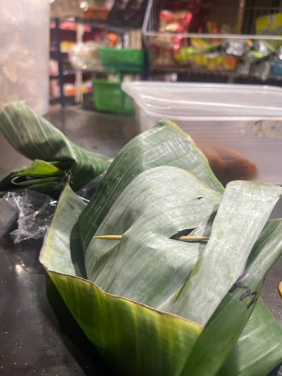 The image shows food wrapped in banana leaves on a dark surface. The banana leaves are large and green, with visible veins. One leaf is folded to form a container, holding the food. A toothpick is stuck into one of the leaves. In the background, shelves stocked with various items are visible, suggesting a market or store setting. There are also plastic containers and bags. The lighting is somewhat dim, creating shadows. The location of Barat Yaman, Indonesia is not depicted in this image; it appears to be an indoor setting, possibly a kitchen or market stall. There are no people visible in the image.