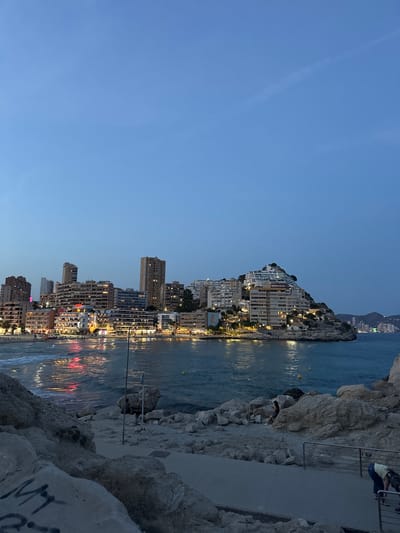 The image depicts a twilight scene of the coastline in La Cala, Spain, likely Benidorm given the architecture. In the foreground, a rocky, uneven terrain slopes down towards the water. Some large rocks are scattered across the area, with a paved path visible running diagonally from the bottom right corner. A metal railing lines the path in the lower right. Further down, closer to the water, there is a sandy area with more rocks. A person is seated on the rocks in the mid-ground, and another person is bent over near a dog on the paved path in the lower right.

The middle ground features the ocean, with gentle waves lapping at the shore. Reflections of lights from the city twinkle on the water's surface. Beyond the water is a densely built-up coastline with numerous tall buildings, many illuminated with lights. The buildings are clustered along the shore and ascend a hill on the right side of the frame, creating a distinct urban landscape against the darkening sky. In the distance, more buildings and possibly mountains are visible on the horizon. The sky is a deep blue, transitioning from a lighter shade at the horizon to a darker hue at the top, with a faint contrail visible. The overall impression is one of a coastal city at dusk, with the lights of buildings and the sea creating a vibrant atmosphere.