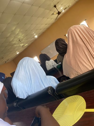 In an indoor setting in Sabo, Nigeria, a view angled upwards captures several seated individuals. One person wears a light pink garment. Another wears a light blue shirt, while a third is dressed in a black and white patterned outfit with a black hijab.

A yellow hand fan rests on a dark wooden table near a black leather chair. The wall features tiles. Overhead, a ceiling fan is visible.