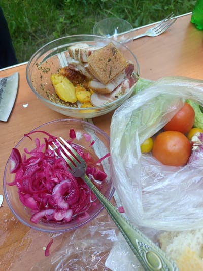 The image displays a rustic outdoor setting, likely a picnic or meal outdoors, with food laid out on a wooden surface. In the foreground, a clear bowl contains sliced red onions that appear to be pickled or marinated, with a fork resting inside. To the right of this, a transparent plastic bag holds various vegetables, including two ripe tomatoes, what appear to be cucumbers, a head of garlic, and a couple of small yellow fruits, possibly lemons or small tomatoes. Behind these items, slices of bread are visible. In the background, another clear bowl contains roasted potatoes and slices of what looks like cured meat or bacon, seasoned with herbs. A knife is positioned to the left of the bowls, and a fork is visible on the table behind the first bowl. The environment suggests a natural setting with greenery visible in the upper background, and the wooden surface indicates a table. The overall impression is one of a casual, freshly prepared meal. Given the context provided of the city of Giaginskaya, Russia, this scene likely depicts a common outdoor dining practice in a rural or suburban area of the region.