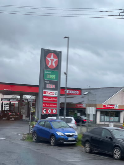 This media file captures a scene at a petrol station on a cloudy day, possibly in Ireland given the currency symbol and language cues. The foreground features a blue car parked, with another black car behind it. A prominent Texaco sign displays fuel prices: "Unleaded 175.9" and "Diesel 175.9". Adjacent to the Texaco signage is a SPAR store, also advertising "Hot Food / Deli" and "Car Wash / NCT Wash". The SPAR sign is clearly visible, as is a blurred "TEXACO" sign on the building. The sky is overcast, suggesting a potentially wet or dull day, and the ground appears wet, indicating recent rain. The overall impression is of a typical, everyday scene at a roadside service station. There are no people clearly visible in the image. The time of day is not explicitly indicated, but the lighting suggests it is daytime.