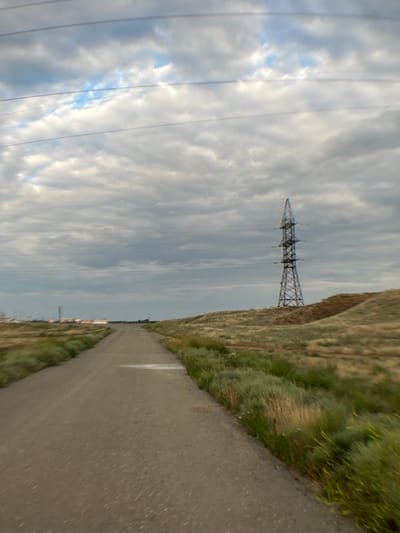 The image depicts a desolate, rural landscape under a cloudy sky. A dirt road stretches into the distance, flanked by dry, grassy terrain. In the middle ground, a tall, skeletal metal pylon stands prominently on a rise, indicating the presence of power lines. In the far distance, a cluster of industrial structures with illuminated lights can be faintly seen, suggesting a town or facility on the horizon. The overall impression is one of isolation and a somewhat industrial yet natural environment. The specific location is identified as the city of Ilyinka, Russia.