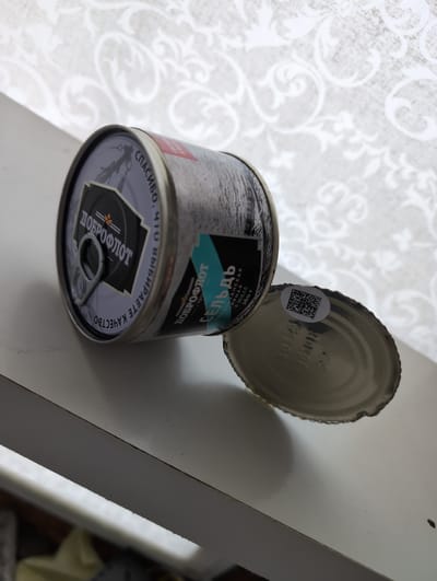 A slightly angled, close-up, eye-level shot shows an opened can of fish. The can is primarily silver and grey, with a label that features the brand "ДОБРОФЛОТ" in large, bold lettering at the top. Below this, there is text that is partially obscured. A red rectangle with white text is visible near the top of the can. The can is lying on its side on a white surface, possibly a shelf. The lid of the can is detached and rests next to it, also on the white surface. The inside of the lid has a circular white sticker with a QR code printed on it. There is also some embossed text on the inside of the lid that is difficult to read. The background consists of a white, textured wallpaper with a repeating floral pattern. No people are present in the image. The scene appears to be indoors. The location of Вышки, Russia is not depicted in the image.