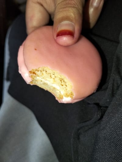 A hand holds a partially eaten pink cookie. The cookie has a bite taken out, revealing a white cream filling and a textured cookie interior. The hand's fingernail is visible and displays a dark red discoloration near the cuticle. The background shows a blend of gray and black fabric, likely clothing. The image is somewhat dimly lit.