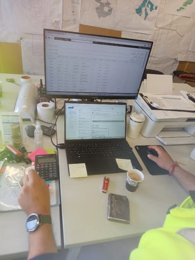 The scene depicts an active office workstation in Drugan, Bulgaria. Two light-colored desks are arranged against a wall covered with numerous paper maps and diagrams. The area is brightly illuminated, with a visible yellowish light source originating from the left.

On the left side of the workstation, a person's left arm, wearing a dark-strapped smartwatch, holds a small, light-colored circular object above a black calculator positioned on a document displaying a map pattern. Nearby items include a green permanent marker, a pink sticky note, a small silver container, a white cylindrical appliance, green-handled scissors, a roll of white paper, a clear bottle of hand sanitizer, a black computer mouse, a calendar, and a multi-layered clear container holding different colored materials.

On the right side of the workstation, a person's right arm, clad in a neon yellow garment and featuring a red string bracelet, operates a black computer mouse on a black mousepad. An open black laptop displaying an email client is positioned next to the mousepad, with two yellow sticky notes affixed to its palm rest. Also on the desk are a red lighter with a Ferrari logo, a dark metallic case with an etched design, and a black and white disposable coffee cup containing a beverage.

A large black desktop monitor is prominently displayed behind the laptop, exhibiting a tabular dataset or spreadsheet interface. To the right of the laptop, a white multi-function printer with papers and a clipboard resting on its top surface is present. A white travel mug is positioned next to the printer.