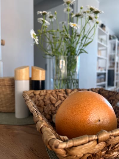 A grapefruit rests in a woven basket on a wooden surface. Behind the basket, two cylindrical objects with wooden tops are visible, one white and one black. In the background, a bouquet of daisies in a glass vase is slightly out of focus. Further in the background, a white bookshelf filled with items is visible. The scene appears to be indoors, likely a domestic setting. There is no indication of the city of Saburovo, Russia in the image.