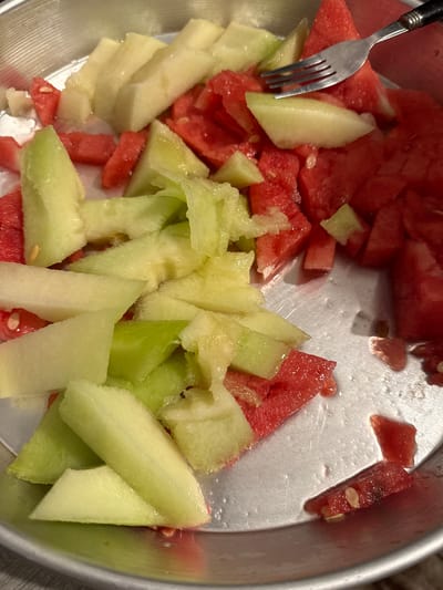 A close-up, overhead shot displays a metal bowl filled with chopped watermelon and cantaloupe. The watermelon pieces are vibrant red, some with visible seeds, while the cantaloupe chunks are a pale green and yellow. A silver fork is partially submerged in the fruit, with its tines piercing several pieces. The bowl appears to be sitting on a surface that could be a table or countertop. The lighting suggests an indoor setting, possibly during the day. There is no visible text in the image. The scene is casual and appears to be set in a domestic environment in Fulda, Germany.