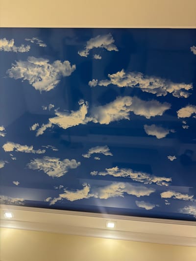 The image shows a ceiling with a sky and clouds mural. The mural is bright blue with white and gray clouds of various sizes and shapes. The ceiling is framed by a white border with recessed square lights. The walls below the ceiling are a light yellow or beige color. There are no people or animals visible in the image. The image does not provide any information about the location in the city of Goris, Armenia.