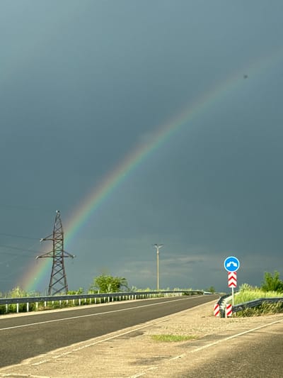 An overcast sky with a prominent rainbow arches over a highway in Karachunivka, Ukraine. To the left, a tall, latticed metal power pylon stands against the dark clouds. The highway stretches into the distance, bordered by a metal guardrail and a grassy verge. On the right side of the road, a traffic sign indicates a double curve ahead, accompanied by striped markers. A streetlamp is visible in the mid-ground.