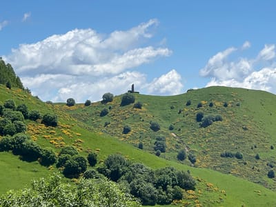The image shows a lush green hillside in Kumlistsikhe, Georgia, under a bright blue sky with scattered white clouds. A solitary, dark, pillar-like monument stands atop the highest peak of the hill. The hillside is dotted with scattered trees and patches of vibrant yellow flowers, adding a touch of color to the verdant landscape. In the foreground, a dense cluster of green foliage suggests the presence of more trees. The overall scene depicts a serene and natural environment, with the monument serving as a focal point against the backdrop of rolling hills.
