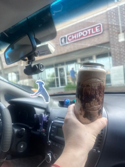 The image shows a hand holding a tall glass filled with a brown beverage, possibly a milkshake or smoothie with chocolate syrup drizzled inside. The glass has some embossed lettering, which is partially visible. The background suggests the setting is inside a car. Through the windshield, a Chipotle restaurant sign is clearly visible, indicating a location in Glenview, United States of America. The interior of the car shows a dashboard with a rearview mirror, a dashcam mounted above it, and an air freshener in the shape of a blue dog hanging from the mirror. The car's steering wheel and part of the center console are also visible.
