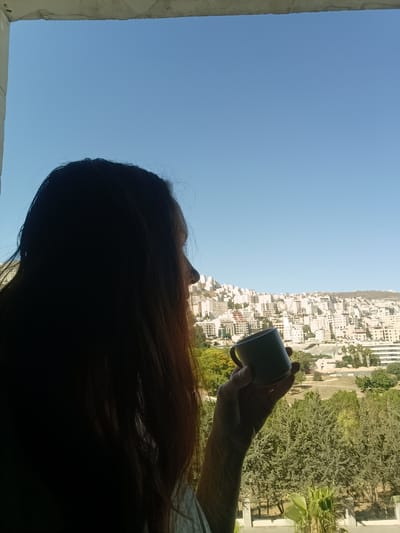A silhouette of a person with long, dark hair is shown from the side, holding a dark mug up to their face. The person is positioned in the foreground, obscuring their features. Behind them, a clear blue sky stretches over a dense city landscape. Buildings with light-colored facades are tightly packed together, climbing up a hilly terrain. Green trees and vegetation are visible interspersed throughout the city and in the foreground, suggesting an outdoor setting within an urban environment. The overall impression is that of a peaceful moment captured from a vantage point overlooking the city, possibly a residential area in Nablus, Palestinian Territories, given the architectural style and terrain.