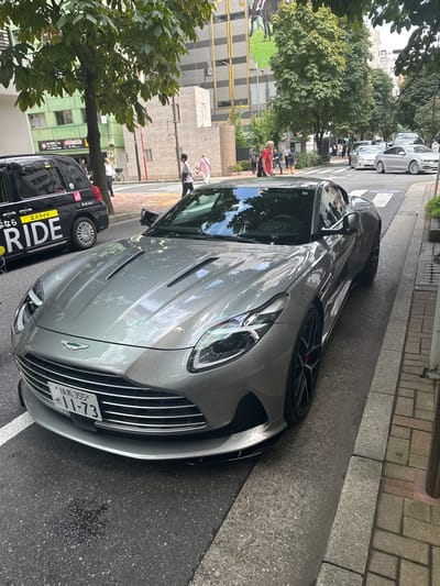 The image depicts a silver-gray Aston Martin DB12 coupe parked curbside on an asphalt street in Chūō, Japan. The vehicle is angled slightly towards the right, showcasing its front grille with horizontal chrome slats, the Aston Martin badge, and a Japanese license plate "練馬 355 せ 11-73". Black multi-spoke wheels with visible red brake calipers are present on the right side. To the vehicle's left and rear, a black S.RIDE taxi is partially visible. In the background, multiple pedestrians traverse sidewalks and a visible crosswalk. The street is flanked by multi-story urban buildings; one prominent structure features a large billboard depicting a horse and jockey. Numerous green-leafed trees line the street, casting reflections on the car's paintwork and windshield. A small dark liquid stain is visible on the asphalt in front of the DB12's bumper.