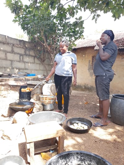 The image depicts an outdoor cooking scene in Vodni, Nigeria, featuring two adult females preparing and consuming food under daylight conditions.

The woman on the left, wearing a light grey polo shirt and dark pants, is actively engaged in cooking. She stands over a makeshift outdoor stove where a dark, shallow pan rests on rocks, heated by an unseen fire from below. She holds a slotted spoon containing multiple fried dough balls, from which oil is dripping, indicating they have just been removed from the hot oil. Her head is turned slightly to the right, and she is smiling. To her right, a woven basket elevated on a metallic stand holds additional fried dough balls, with a metal pot positioned beneath it.

The woman on the right, dressed in a dark polo shirt, dark shorts, and a black patterned headscarf, stands slightly behind and to the right of the first woman. She is actively eating, holding a piece of food to her mouth with her right hand. Her left hand is near her stomach, and she is also smiling or laughing.

In the foreground, on the reddish-brown soil ground, there are several dark, circular basins. One basin in the center contains multiple un-fried dough balls. A small, light-colored wooden stool is positioned to the left foreground. In the mid-ground, two large, dark, cylindrical pots or drums are visible.

The background includes a low, unplastered concrete block wall behind the cooking area. Beyond the wall and to the right, a building with a corrugated metal roof and unpainted walls is visible. A large tree with green foliage extends into the upper left and center of the frame, providing some overhead cover.