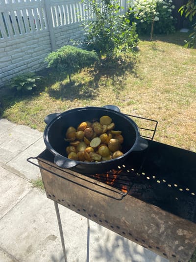 This media file shows a top-down view of a large, black, cast-iron pot filled with roasted potatoes. The pot is resting on a wire rack inside a metal barbecue grill. The grill contains glowing embers and flames, indicating that the potatoes are actively being cooked. The scene is set outdoors in a grassy yard with a concrete patio area. In the background, a white picket fence, some green bushes, and trees are visible. The lighting suggests it is daytime, likely sunny, given the bright highlights and shadows. The overall atmosphere appears to be relaxed and casual, possibly a backyard gathering or a picnic. There is no visible text in the image. The location context is Знаменскі сельскі Савет, Belarus.