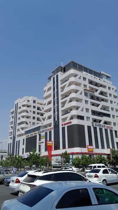 The image shows a modern white building with a distinctive architectural design, characterized by layered balconies and angular protrusions, under a clear blue sky. The building appears to be a mixed-use commercial and residential complex. On the lower levels, there are prominent signs for "EMIRAN BAZAR" and "DON ARCHI," indicating a marketplace and a restaurant or food establishment. Trees line the street in front of the building. Several cars are parked or driving on the street in the foreground, including a white sedan, a silver sedan, and a light blue sedan. People are visible walking on the sidewalk. The scene is likely a street view in the city of Denov, Uzbekistan, given the building's signage and the overall urban environment.