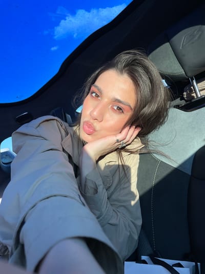 A close-up, high-angle selfie captures a young woman inside a car, looking directly at the camera with a playful "kiss" face. Her lips are puckered, and her left hand is raised to her cheek, displaying several silver rings, including a star-shaped one. She has dark brown hair and is wearing a light-colored (beige or khaki) button-up shirt or jacket. Her makeup features prominent pink/coral eyeshadow and glossy lips.
The setting is the interior of a car, with sunlight streaming in and casting distinct shadows. The car seat behind her is black and grey with light green stitching and a stylized logo, possibly a winged 'A' or 'V', embossed on the headrest. Through the windshield on the left side of the frame, a clear blue sky with some scattered white clouds is visible, indicating daytime and good weather. Small water droplets or dust are present on the windshield. A white shopping bag with black handles is partially visible on the right side of the image, next to the woman. The overall impression is bright and casual. The scene takes place in Apaga, Armenia. No discernible text is visible.