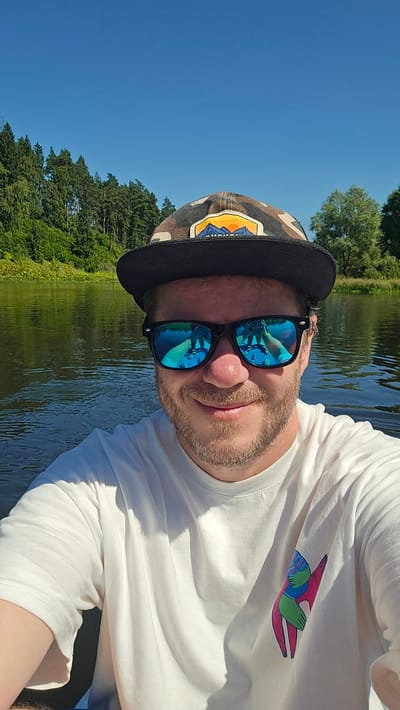A man with a beard and sunglasses is taking a selfie on a body of water, likely a river or lake, on a sunny day. He is wearing a white t-shirt with a colorful graphic design on the left side. He is also wearing a camouflage baseball cap with an embroidered patch featuring mountains and a sunset. The reflection in his sunglasses shows another person and possibly a kayak or raft. The background consists of trees and a clear blue sky. The location is Voinskaya Chast' Mchs, Russia.