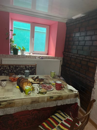 A table is set with food and drinks in a room with pink walls and a brick stove. On the table, there is a loaf of bread, sliced bread, a plate of sliced sausage, several cups, and a can. A window with green panes is visible behind the table. A radiator is located beneath the window. A chair with a colorful knitted seat cover is pulled up to the table. The overall impression is a simple meal in a rustic setting.