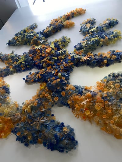 The image shows a detailed, three-dimensional art installation mounted on a white wall. The artwork is composed of numerous small, organic-shaped elements in shades of blue, orange, and yellow, resembling coral or marine life. These elements are densely clustered and appear to be made of translucent or semi-translucent materials, possibly glass or resin, with some appearing to have a lace-like texture. The installation is arranged in abstract, flowing forms that extend across the wall, with some sections appearing more layered and volumetric than others. The lighting casts subtle shadows, highlighting the textured surface of the artwork and the white wall. The overall impression is one of an intricate, underwater ecosystem brought to life. This artwork is located in the lobby of the Inter-American Development Bank, 1300 New York Avenue NW, Washington, D.C. 20577, United States of America.