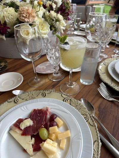 The image shows a table setting, likely for a meal, with an assortment of items.
**Objects:**
* Several drinking glasses, some filled with clear liquid, and one with a yellow beverage garnished with a blackberry and mint.
* Plates with cheese, grapes, and charcuterie (prosciutto).
* Cutlery: forks, spoons.
* Floral arrangement with roses and other flowers.
* Charger plates with ornate edges.
**Environment:**
* The table appears to be made of wood.
* The setting suggests a formal or semi-formal dining occasion.
It is impossible to determine if this is the city of Orizaba, Mexico.