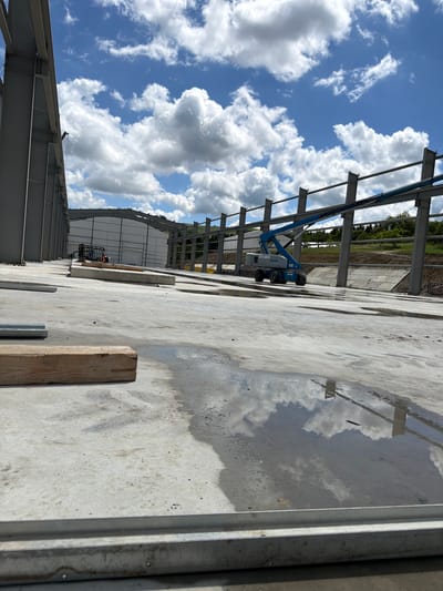 The image depicts a construction site in Skrzydlna, Poland, under a cloudy sky. The foreground is dominated by a concrete surface, with puddles of water reflecting the sky and surrounding structures. A wooden beam lies horizontally in the middle ground, to the left. In the background, a large steel-framed building under construction is visible, with a blue aerial work platform positioned alongside it. Further in the distance, more construction elements and a hilly landscape with trees can be observed. The overall scene suggests ongoing construction work in an outdoor setting.