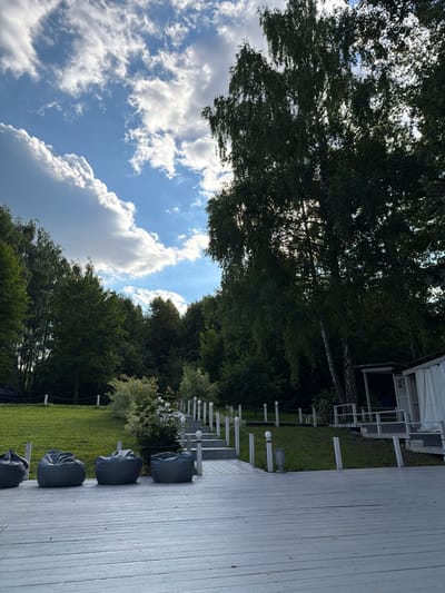 The image depicts an outdoor scene in Povedniki, Russia, featuring a wooden deck in the foreground with four dark grey beanbag chairs arranged along its edge. Beyond the deck, a pathway with white railings and lights leads uphill through a grassy area dotted with small trees. In the background, dense green foliage of various trees forms a natural backdrop. To the right, a white structure with a covered porch is visible. The sky above is a bright blue with scattered white clouds, and sunlight filters through the leaves of a large tree on the right side of the frame.